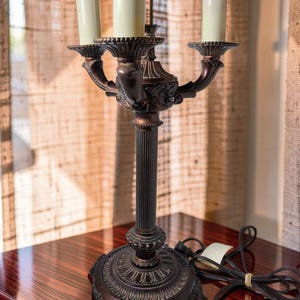 May include: A dark bronze-toned metal candelabra lamp featuring a central fluted column and a decorative circular base with ornate detailing. The lamp holds three off-white cylindrical candle-style bulbs and includes a black electrical cord.