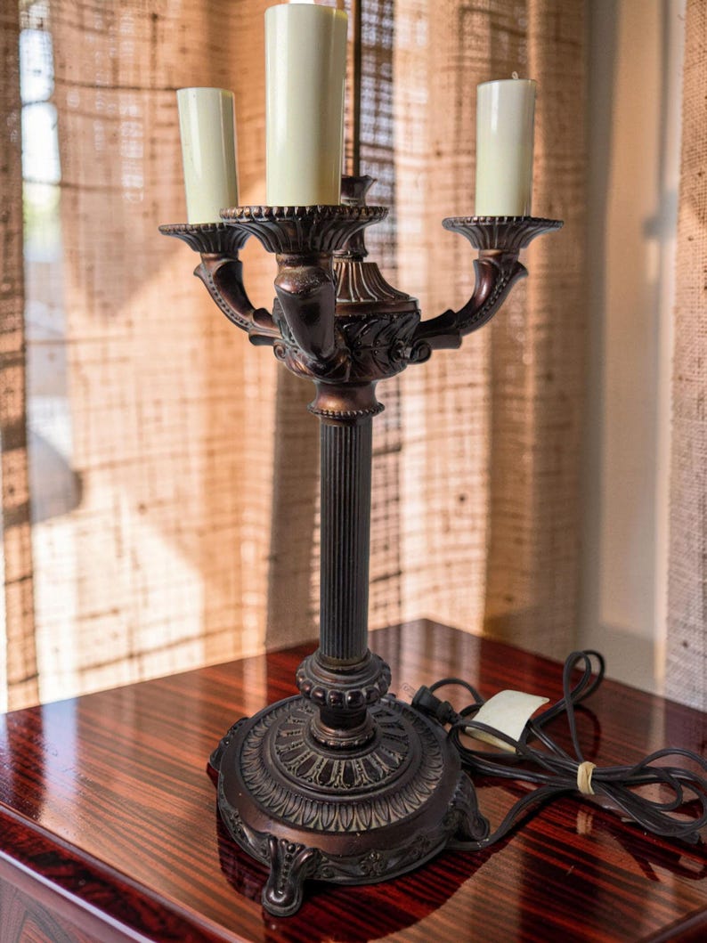 May include: A dark bronze-toned metal candelabra lamp featuring a central fluted column and a decorative circular base with ornate detailing. The lamp holds three off-white cylindrical candle-style bulbs and includes a black electrical cord.