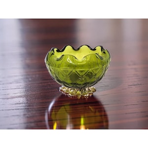 May include: A small, green glass bowl with a ruffled rim and diamond pattern. The bowl sits on a gold-coloured, starburst-shaped base. The bowl's reflection is visible on the dark wooden surface.