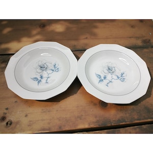 May include: Two white, octagonal-shaped plates with a blue rose design in the center. Each plate has a thin blue line around the rim. The plates are set on a rustic, wooden surface.