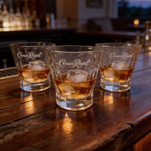 May include: Three clear glass tumblers, each etched with the Crown Royal logo, containing amber-colored liquid and square ice cubes. The glasses feature a faceted, grid-like pattern on the exterior surface and sit on a dark wooden bar top.