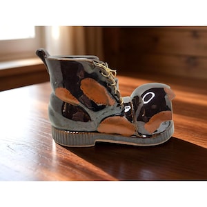 May include: A ceramic boot-shaped planter in shades of brown, tan, and gray. The boot features a textured surface, tan patches, and gold-colored laces. The planter is designed to hold small plants or flowers, adding a unique touch to home decor.