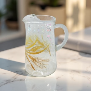 May include: Clear glass pitcher with a handle, featuring a gold feather design. The pitcher has a spout and is filled with glitter. The pitcher is designed for serving beverages.