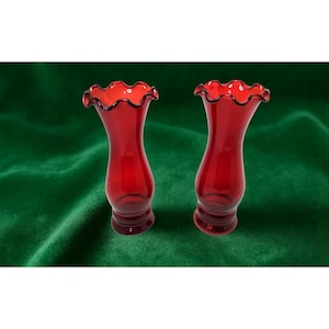 May include: Two ruby red glass vases with ruffled rims and a flared base. The vases have a slender, hourglass shape and are displayed against a green background. The vases are likely for decorative purposes.