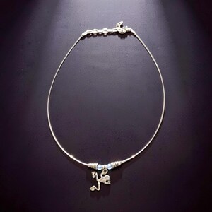 May include: A silver-toned necklace with a delicate wire design. The necklace features a small charm of a stylized figure and two small blue beads. The clasp is a series of small, connected links. The necklace is displayed against a dark background.