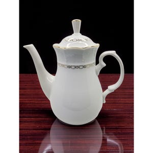 Op de afbeelding: Witte porseleinen theepot met een gebogen tuit en handvat. Het deksel en de body zijn versierd met gouden randen en een delicaat zwart-gouden patroon. De theepot heeft een hoog, slank ontwerp.