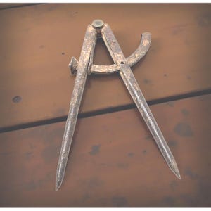 May include: An antique, rusty metal compass with pointed tips, a curved arm, and a central pivot. The compass is set against a brown wooden surface, showcasing its aged appearance and historical significance. This tool is likely used for measuring distances or drawing circles.