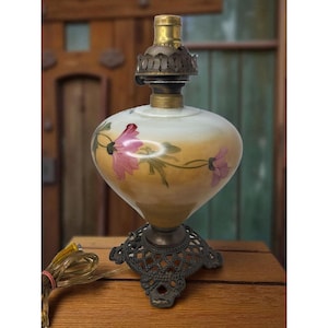 May include: An antique oil lamp with a floral design. The glass globe is painted with pink flowers and green leaves against a yellow and white background. The lamp has a brass top and a decorative metal base.