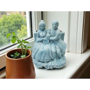 May include: A pale blue ceramic planter depicting a seated couple in historical attire. The planter has two openings for plants and is on a white windowsill. A small terracotta pot with a green plant is to the left of the planter.