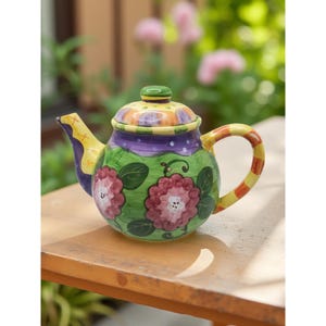 May include: A hand-painted ceramic teapot with a floral design. The teapot features a green body with pink flowers, a purple band, and a yellow and orange lid. The handle is yellow with red stripes. The spout is purple and yellow.