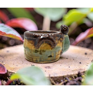 May include: A small, ceramic turtle-shaped planter. The planter has a brown, green, and yellow glaze. The turtle's head forms a handle. The planter is on a wooden surface, surrounded by greenery.