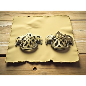 Può includere: Due maniglie antiche in ottone con disegni simmetrici ornati. Le maniglie sono poste su un pezzo di carta pergamena, che poggia su una superficie di legno. Estetica vintage.