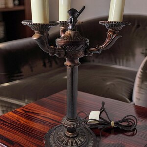 May include: A three-light candelabra lamp with a dark bronze-toned metal finish. The lamp features a fluted central column, a decorative circular base with ornate detailing, and three cream-colored candle-style light covers on curved arms.