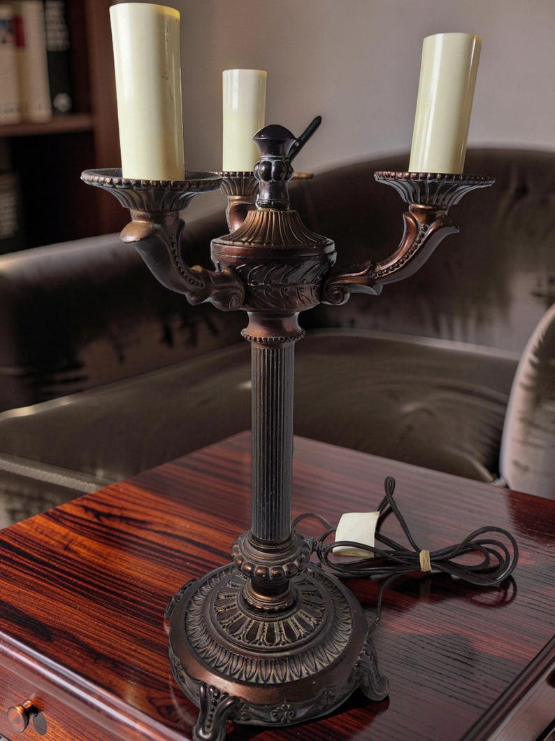 May include: A three-light candelabra lamp with a dark bronze-toned metal finish. The lamp features a fluted central column, a decorative circular base with ornate detailing, and three cream-colored candle-style light covers on curved arms.
