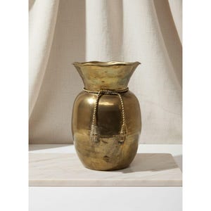 May include: A brass vase with a wide, ruffled rim and a rounded body. A rope with tassels is tied around the neck. The vase sits on a marble surface against a neutral backdrop.