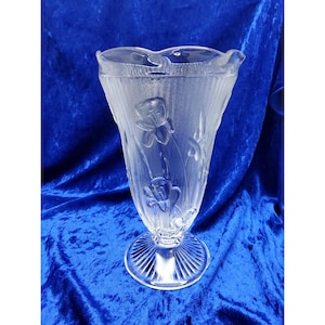 May include: Clear glass vase with a flared top and a pedestal base. The vase features embossed floral designs and vertical textured lines. The vase is approximately 23 cm tall.