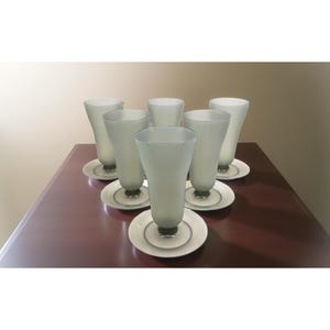 May include: A set of six frosted glass goblets with matching saucers. The goblets have a tall, slender shape and sit on small, round saucers. The glassware is arranged on a dark brown surface, creating a classic and elegant aesthetic.