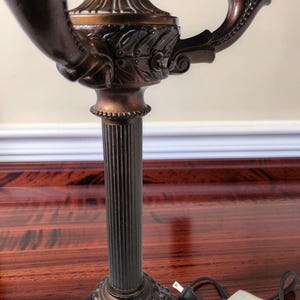 May include: A bronze-toned metal candelabra lamp featuring a central fluted column, a circular base with ornate detailing, and two curved arms extending outward. The lamp includes a brown electrical cord with a standard two-prong plug attached.
