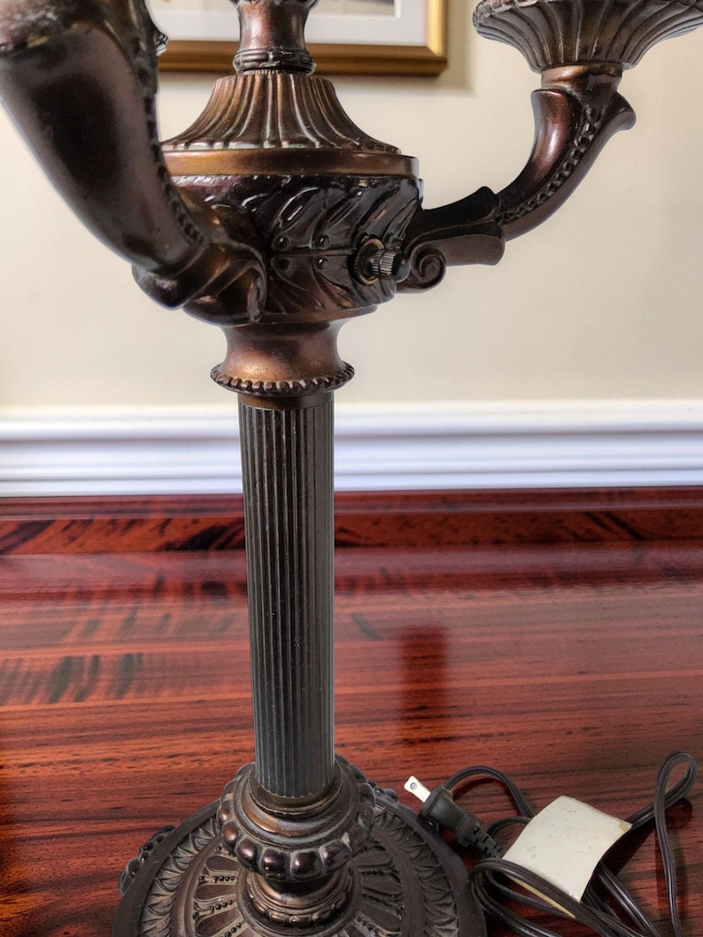 May include: A bronze-toned metal candelabra lamp featuring a central fluted column, a circular base with ornate detailing, and two curved arms extending outward. The lamp includes a brown electrical cord with a standard two-prong plug attached.
