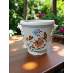 Op de afbeelding: Witte keramische bloempot met twee handvatten en een bloemenontwerp in tinten oranje, blauw en bruin. De bloempot heeft een ronde basis en is geschikt voor binnen of buiten.