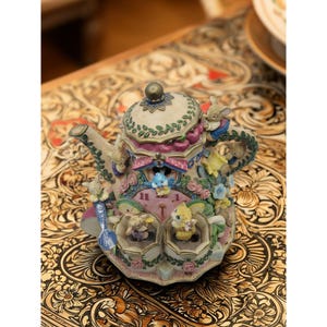 May include: A decorative teapot with a whimsical design featuring small animal figurines, flowers, and a clock face. The teapot is painted in pastel colors with intricate details, including a handle and spout.
