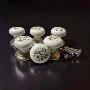 May include: A set of six white ceramic cabinet knobs with a floral pattern featuring green leaves and white flowers. Each knob has a metallic base and is accompanied by several silver-toned metal screws for installation on furniture or cabinetry.