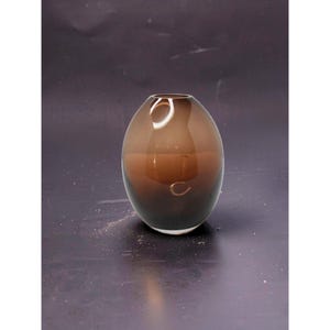 May include: A brown glass vase with an oval shape. The vase has a gradient effect, transitioning from a lighter brown at the top to a darker brown at the base. The vase is transparent, allowing light to pass through.
