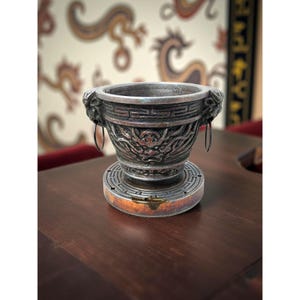 May include: A detailed, silver-toned decorative bowl with dragon and lion head designs. The bowl has a circular base with a geometric pattern and two metal ring handles. The bowl is sitting on a dark brown surface.