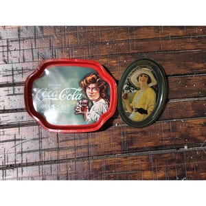 May include: Two vintage Coca-Cola serving trays. One is red and rectangular with a woman holding a glass of Coca-Cola. The other is oval, green, and features a woman in a yellow dress and hat holding a glass of Coca-Cola.