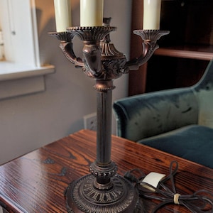 May include: A bronze-toned metal candelabra lamp featuring a central fluted column, a decorative circular base with ornate detailing, and three upright candle-style light sockets with cream-colored covers, accompanied by a black electrical cord.