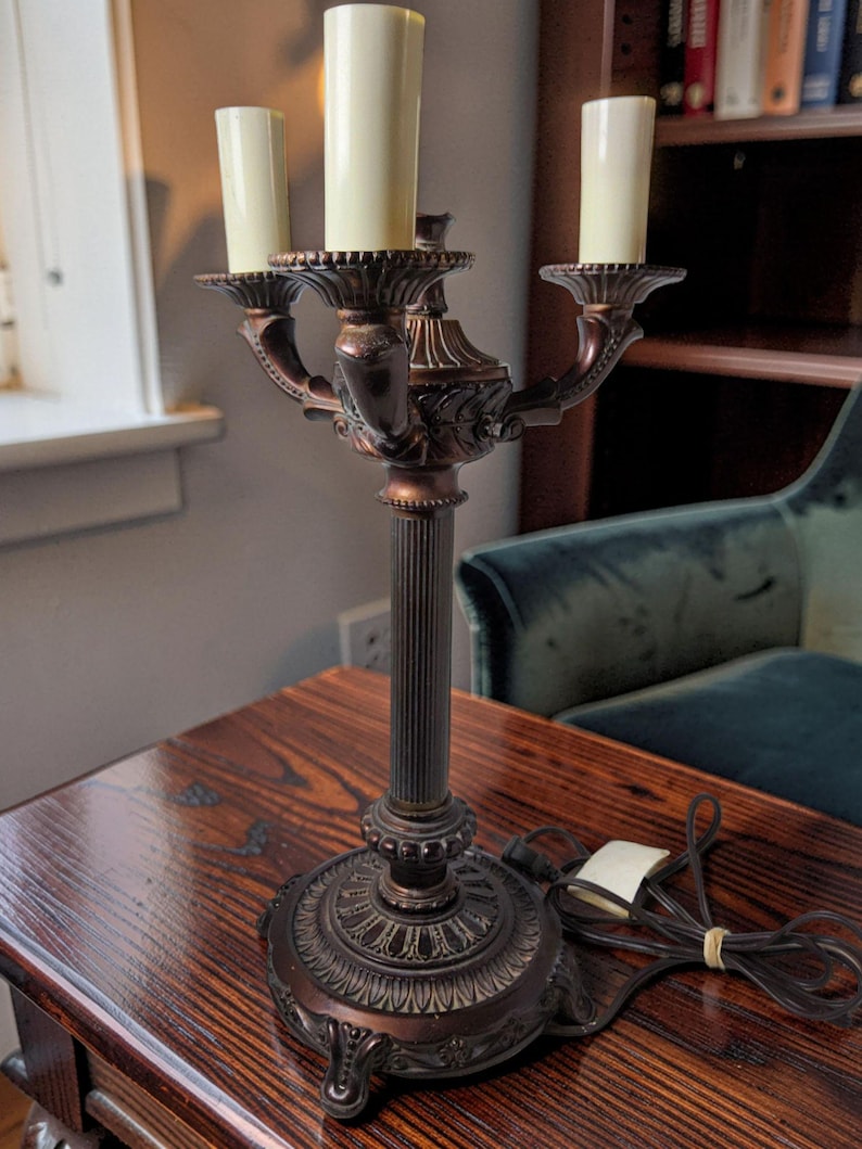 May include: A bronze-toned metal candelabra lamp featuring a central fluted column, a decorative circular base with ornate detailing, and three upright candle-style light sockets with cream-colored covers, accompanied by a black electrical cord.