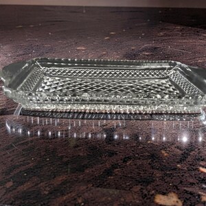 May include: A rectangular, clear glass serving tray with a diamond pattern in the center and a beaded edge. The tray has two curved handles on either end. The tray is sitting on a dark, reflective surface.