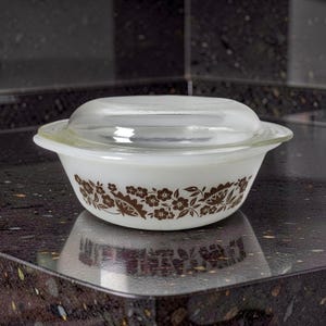 May include: A white glass casserole dish with a clear glass lid. The exterior of the bowl features a repeating brown floral and leaf pattern encircling the sides. The dish has integrated handles on opposite sides and a smooth, rounded base.