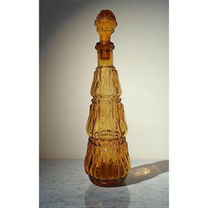 May include: Amber-colored glass decanter with a decorative stopper. The bottle has a ribbed design and a rounded base. The stopper is also amber-colored and has a textured, rounded top. The decanter is likely used for storing and serving beverages.