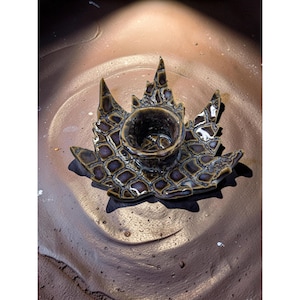 May include: A ceramic candle holder with a dark brown and purple glaze. The holder is shaped like a stylized leaf with a central cup for a candle. The leaf design has a textured, geometric pattern. The candle holder is on a brown surface.