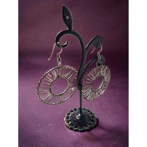 May include: A pair of silver-toned wire earrings with a circular design, displayed on a black metal earring stand. The earrings have a delicate, openwork pattern, and the stand has a decorative base. The background is a solid purple color.