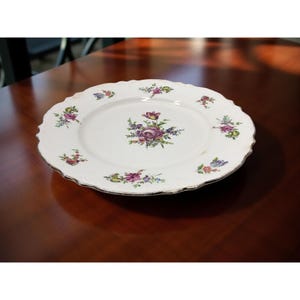 May include: White ceramic plate with a scalloped edge and floral design. The plate features a central floral arrangement and smaller floral clusters around the rim. The flowers are in shades of purple, pink, and green.