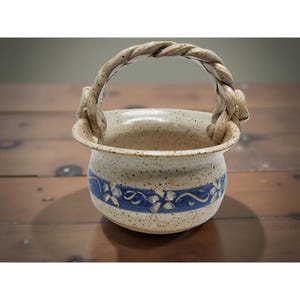 May include: A handmade ceramic basket with a twisted handle. The off-white speckled pot features a blue floral design around the base. The handle is a light tan color. The basket is sitting on a wooden surface.
