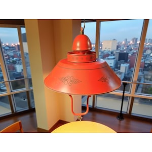 May include: A red metal pendant light with a conical shade and decorative black scrollwork. The light fixture has a bell-shaped top and a curved support. The light is hanging in front of a window with a city view.