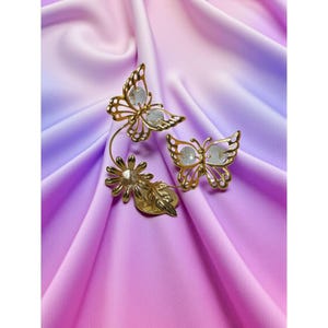 Op de afbeelding: Goudkleurige brocheset met vlinder- en bloemmotieven. De set bevat twee vlinderbroches met transparante centra en een bloem met een centrale edelsteen. De broches zijn verbonden door een gebogen stengel en een blad.