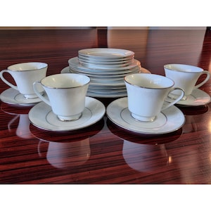 Può includere: Un servizio da tè in porcellana bianca con un delicato motivo floreale e finiture argentate. Il set comprende tazze da tè, piattini e piatti, disposti su una superficie riflettente. Le tazze hanno manici curvi e i piattini un bordo argentato.