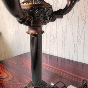 May include: A bronze-toned metal candelabra lamp featuring a fluted central column, a circular base with decorative leaf patterns, and two curved arms extending upward to hold light sockets. A black electrical cord with a plug is attached.