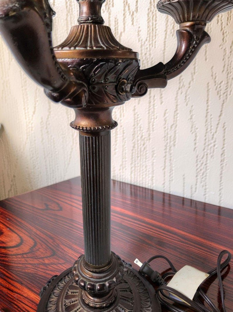 May include: A bronze-toned metal candelabra lamp featuring a fluted central column, a circular base with decorative leaf patterns, and two curved arms extending upward to hold light sockets. A black electrical cord with a plug is attached.