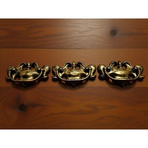 May include: Three antique-style drawer pulls with a gold finish and black accents. The pulls have a decorative, ornate design with a curved handle and detailed embellishments. They are mounted on a wooden surface.