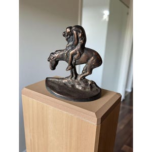Op de afbeelding: Bronzen beeldhouwwerk van een ruiter op een paard. Het beeld is gemonteerd op een lichtbruine houten sokkel. De figuren zijn gedetailleerd, met de ruiter voorovergebogen. Donkere bronzen afwerking.