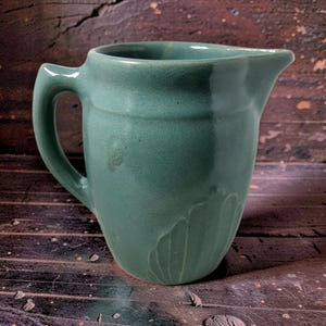 May include: A sage green ceramic pitcher with a glossy finish, featuring a curved handle on the left side and a pouring spout on the right. The lower body of the pitcher displays a subtle, embossed fan-like pattern integrated into the clay surface.