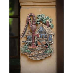 May include: A rustic, three-dimensional wall hanging featuring a water pump, a small house, and a bucket. The piece is painted in shades of red, brown, blue, and green, with a weathered, antique appearance. The house has blue window frames and a green tree.