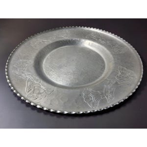 May include: A round, silver-colored metal plate with a hammered texture. The plate features embossed floral designs around the rim and a scalloped edge. The center of the plate is smooth and slightly recessed.
