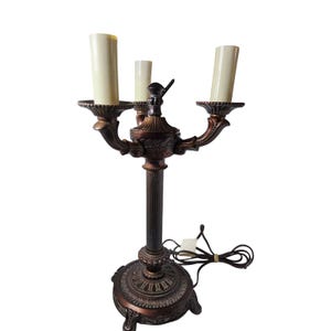 May include: A bronze-toned metal candelabra lamp featuring three upright cream-colored candle sleeves. The lamp has a fluted central column, a decorative circular base with ornate detailing, and a black electrical cord with a switch.