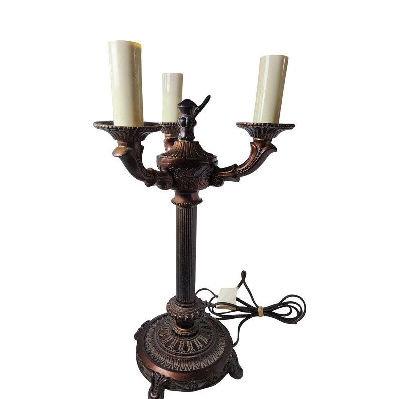 May include: A bronze-toned metal candelabra lamp featuring three upright cream-colored candle sleeves. The lamp has a fluted central column, a decorative circular base with ornate detailing, and a black electrical cord with a switch.